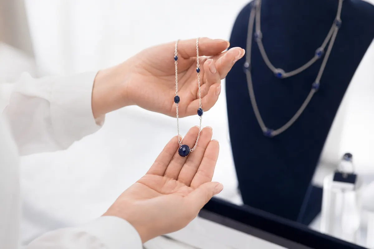 Mãos de uma cliente examinando delicadamente um colar de semijoia em uma loja bem iluminada, com foco nos detalhes da peça.